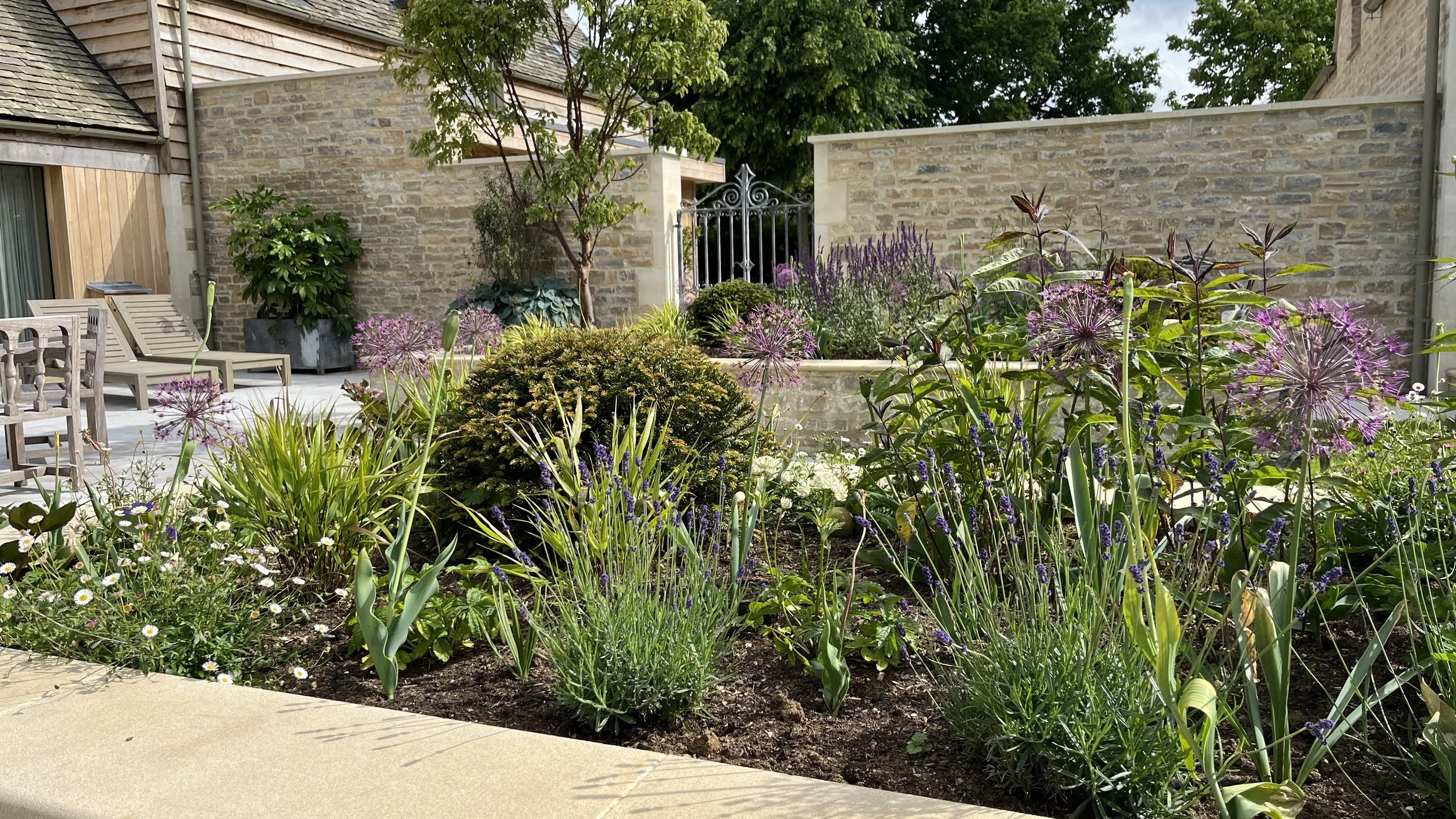 A garden with various purple, white, and green plants and flowers, a stone and brick wall, outdoor furniture, and trees in the background.
