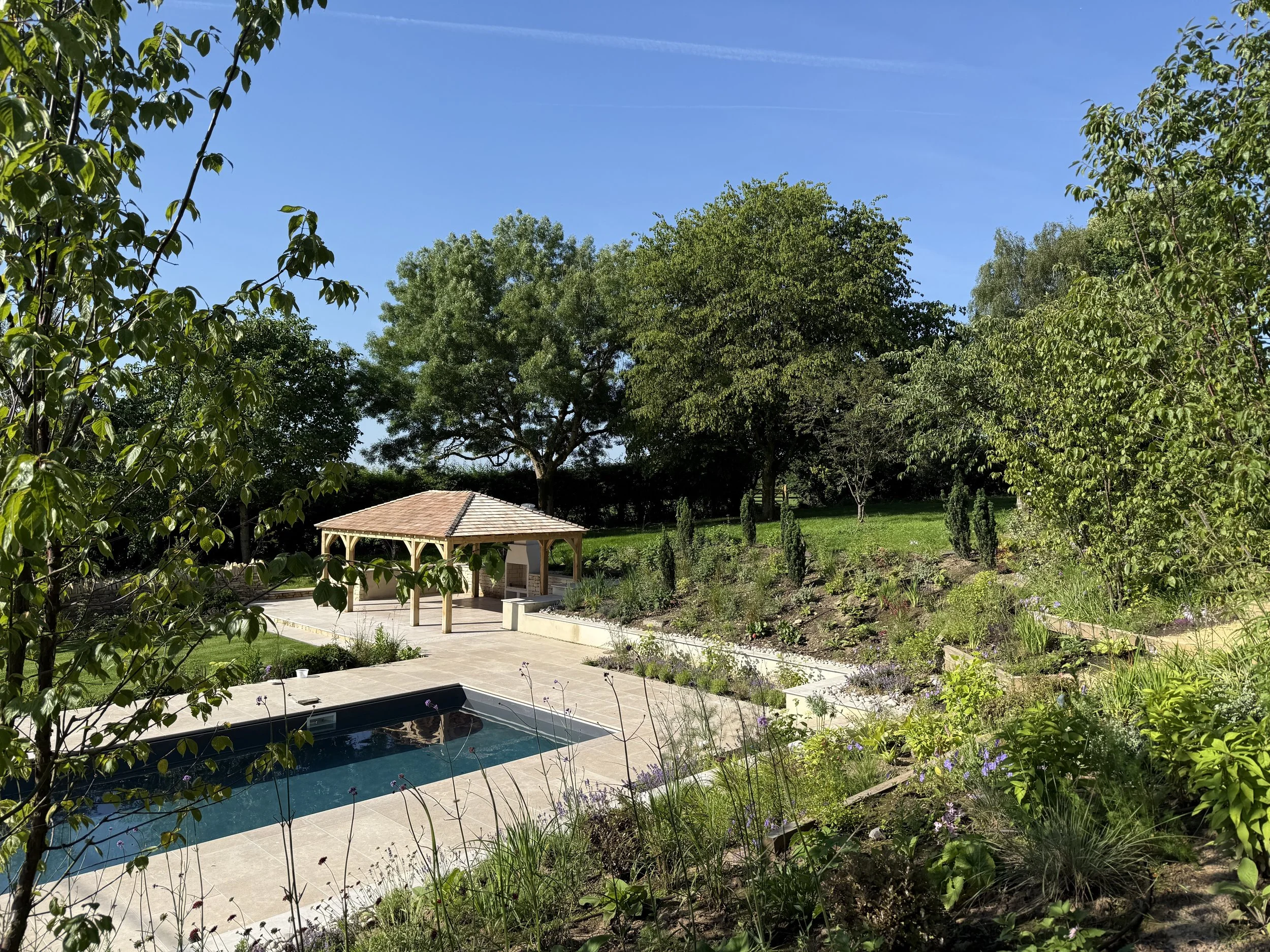 A garden view showing a swimming pool, a shaded pavilion, lush green trees, and a garden with flowers under a clear blue sky.