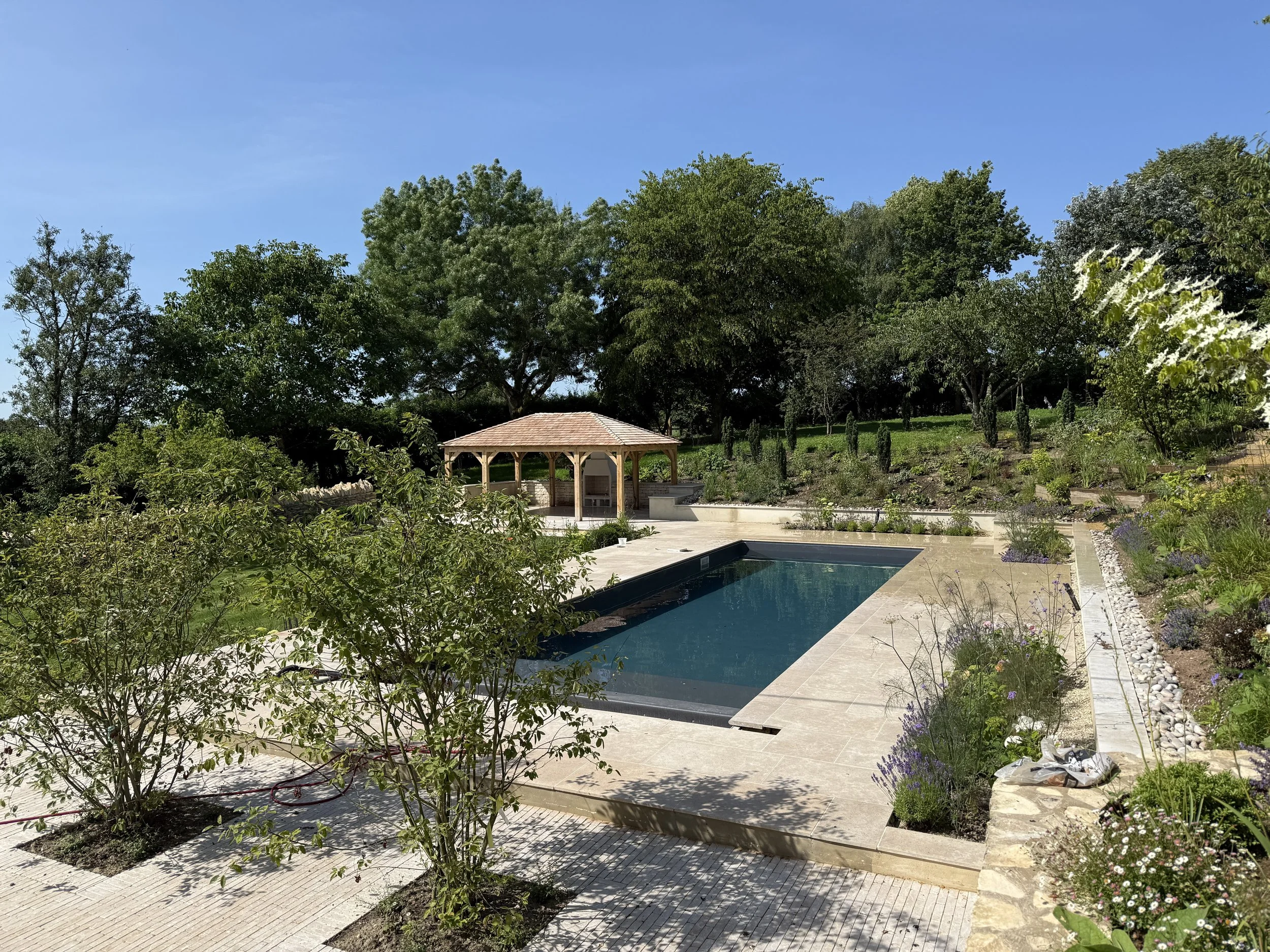 A garden with a swimming pool, a oak gazebo, and trees and plants surrounding the area.