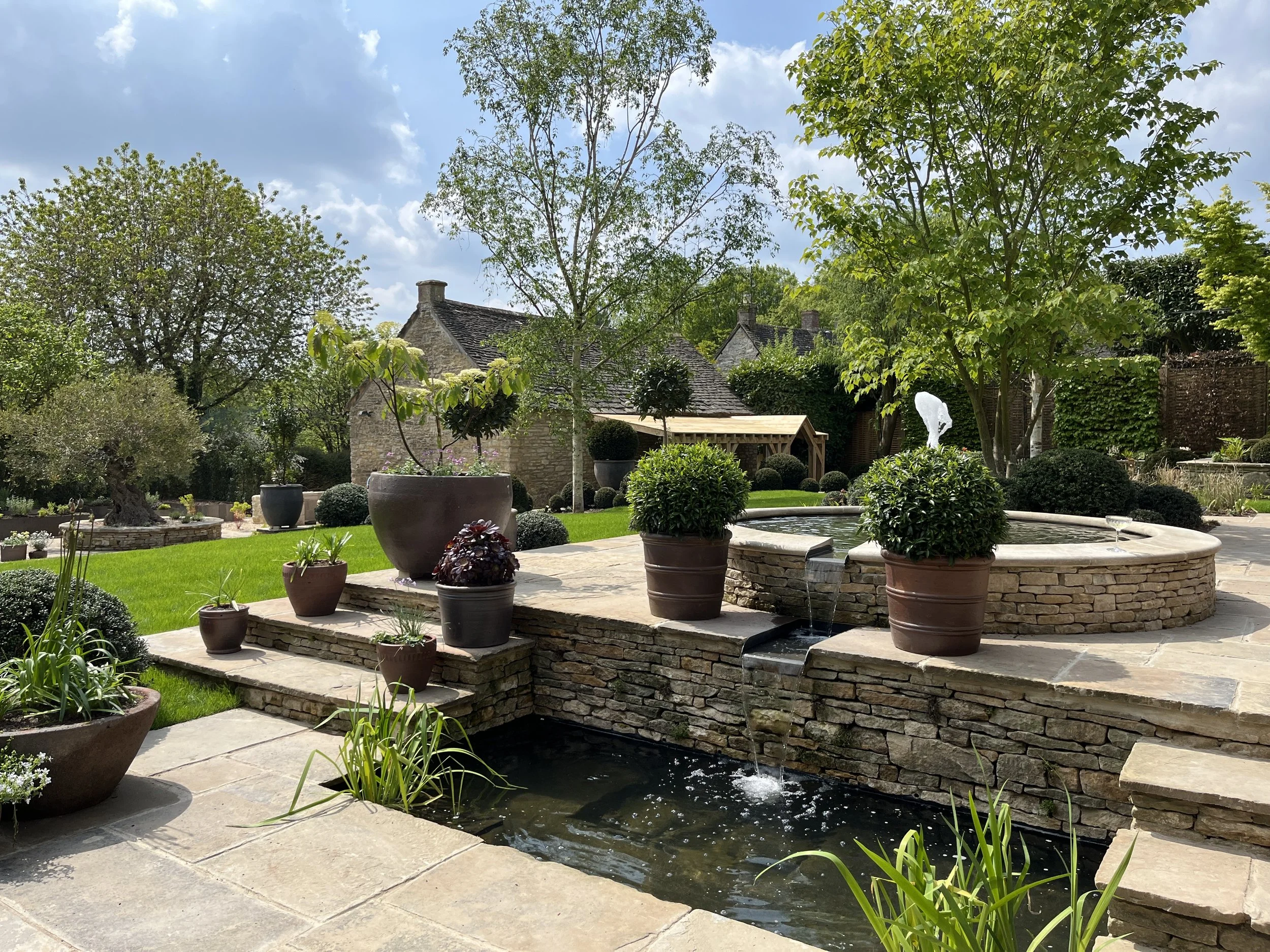 A landscaped backyard with a stone patio, potted plants, a water fountain, lush green grass, trees, and shrubs under a partly cloudy sky.