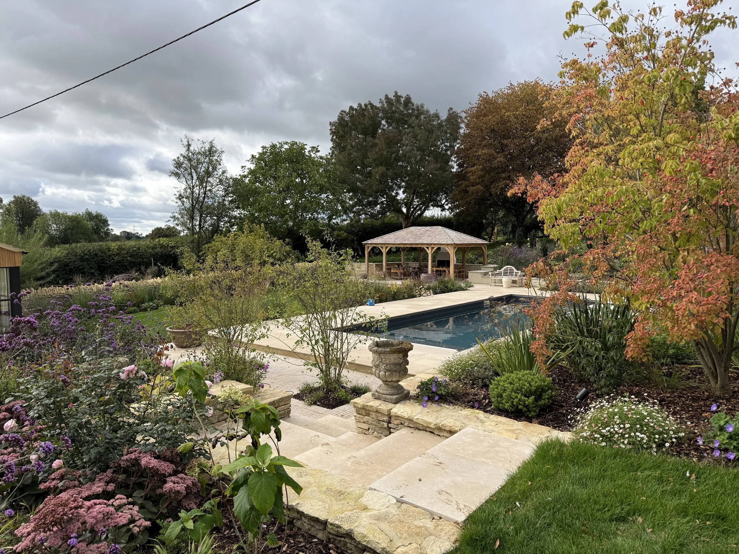 A backyard garden with a swimming pool, surrounded by lush greenery, colorful flowers, and trees, with an overcast sky overhead and a wooden gazebo in the background.