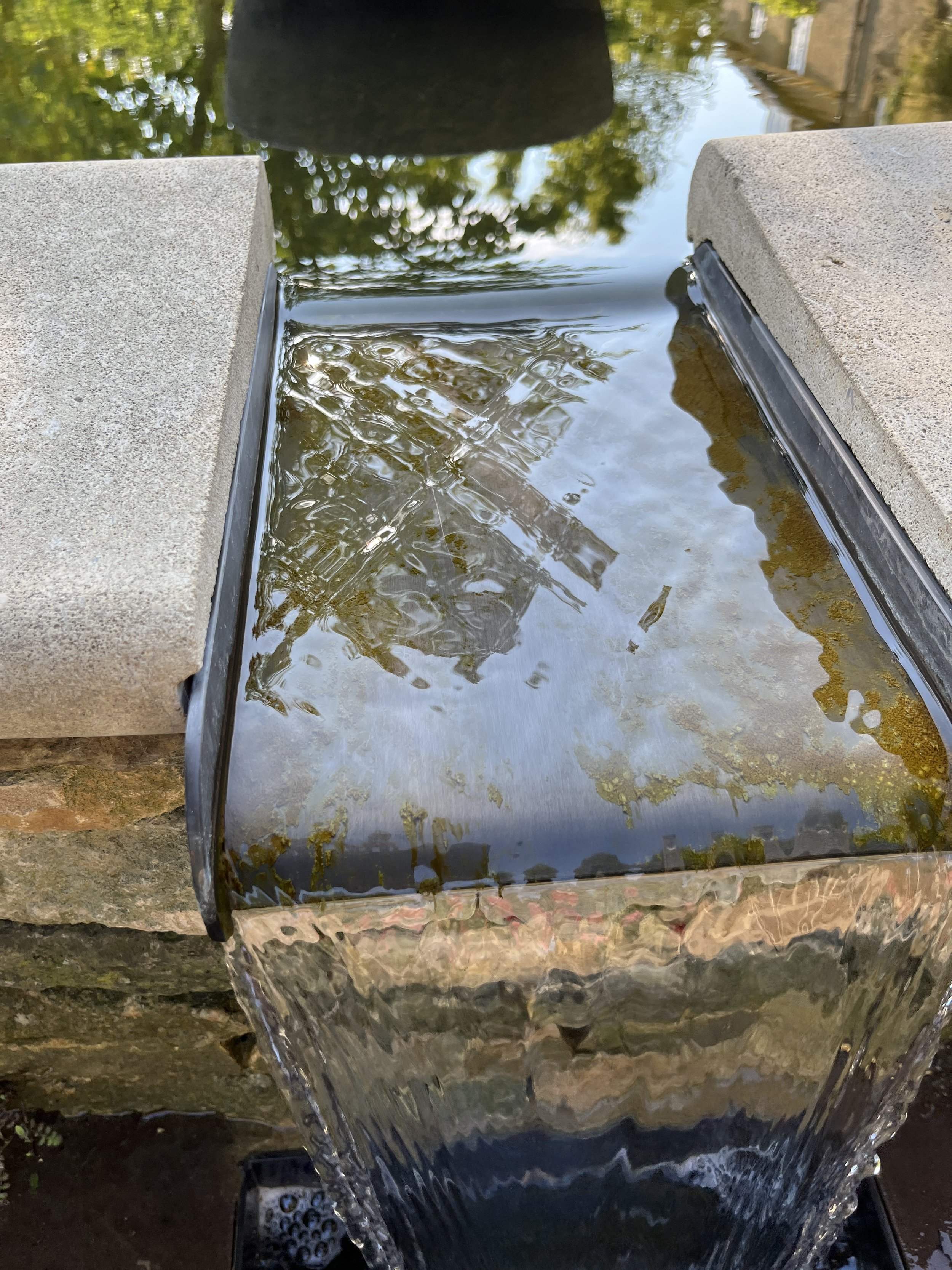 Close-up of a water feature with water flowing over a curved metal structure between two concrete blocks, with reflections of trees and the sky in the water.