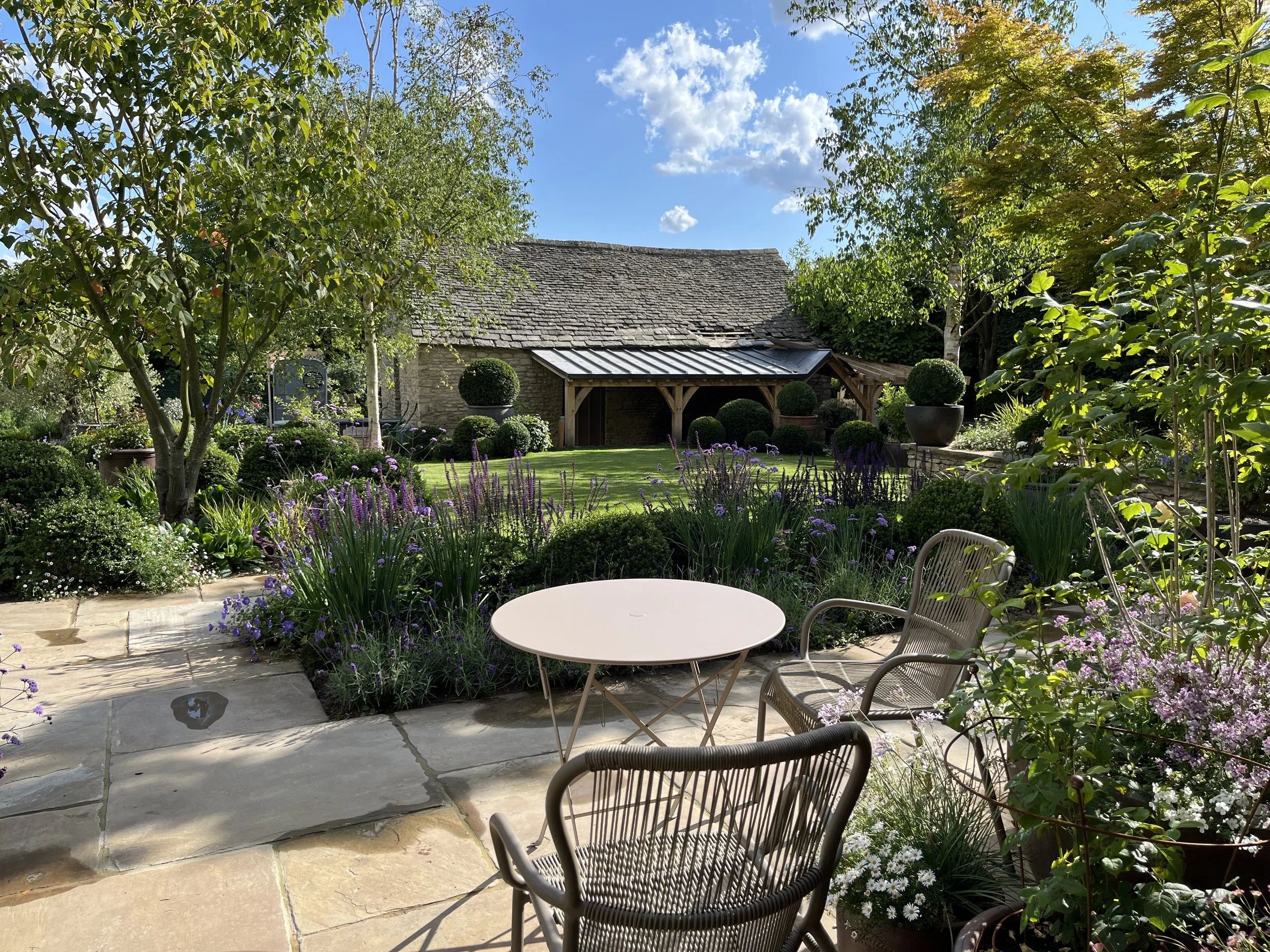A patio with a stone floor, metal chairs, and a round table overlooking a lush garden with various plants, bushes, and trees, and a rustic house in the background under a partly cloudy sky.