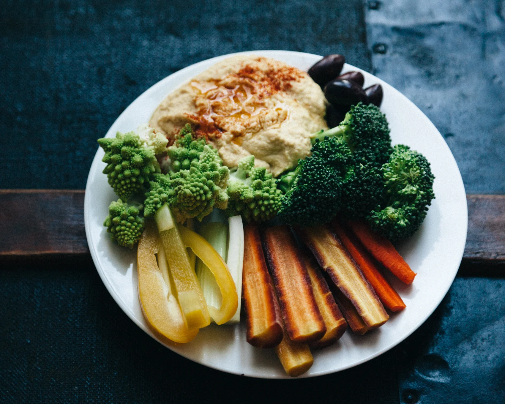 Roasted red pepper hummus garnished with a dollop of olive oil and a sprinkling of paprika, with a side of kalamata olives, romanesco, yellow peppers, celery, carrots and broccoli. 