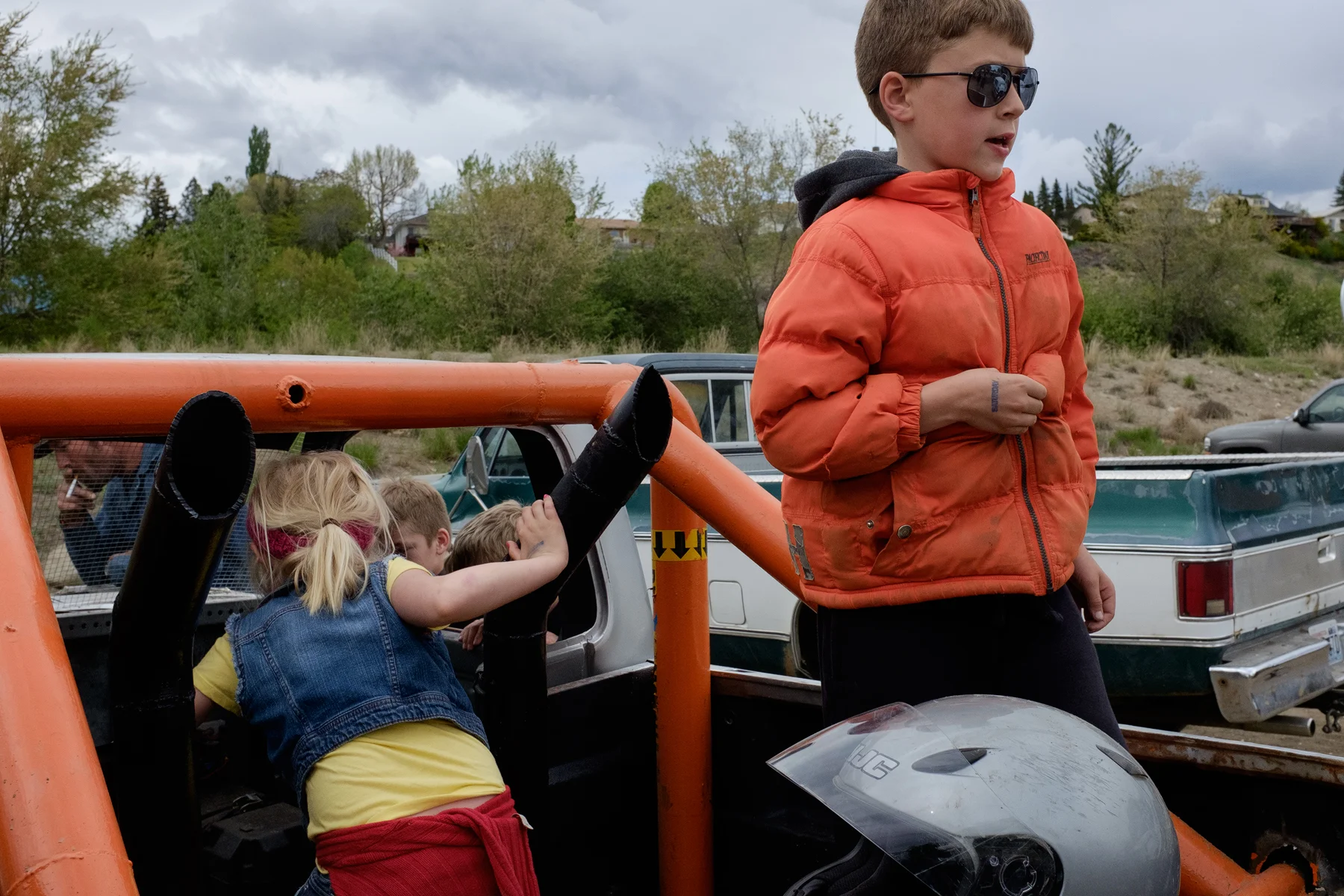 Children stand in the back of a demolition derby car, while dad smokes in the background. Omak, Washington, USA. Image ©Connor Stefanison