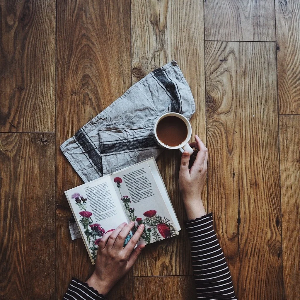 Hands drinking tea and reading wildflower book on wooden table.jpg
