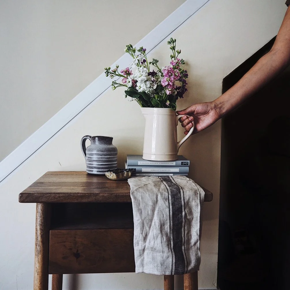 Hand holding a jug of pastel flowers on a pile of books on a side table.jpg