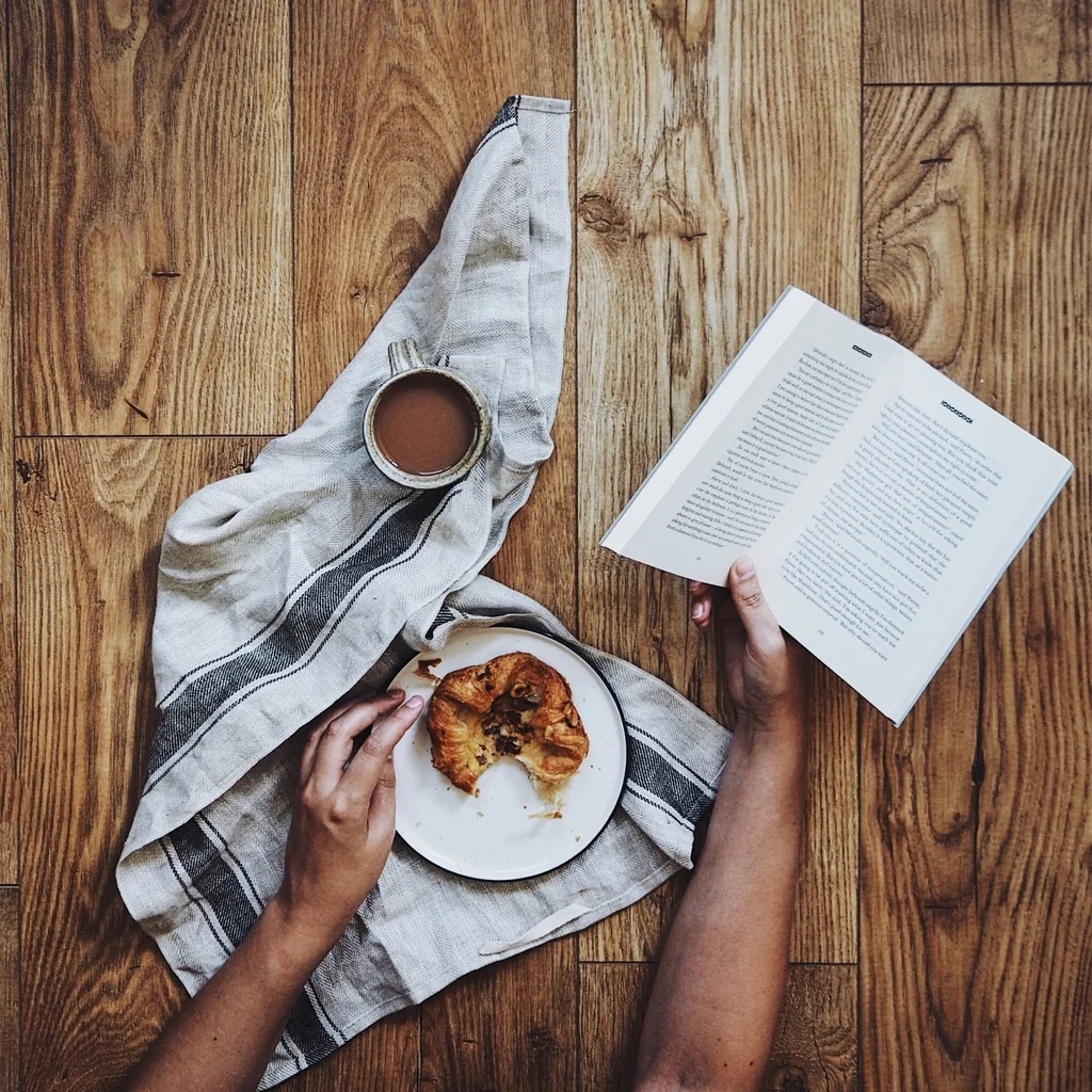 Hands holding a book with a cup of tea and pastry on a table.jpg