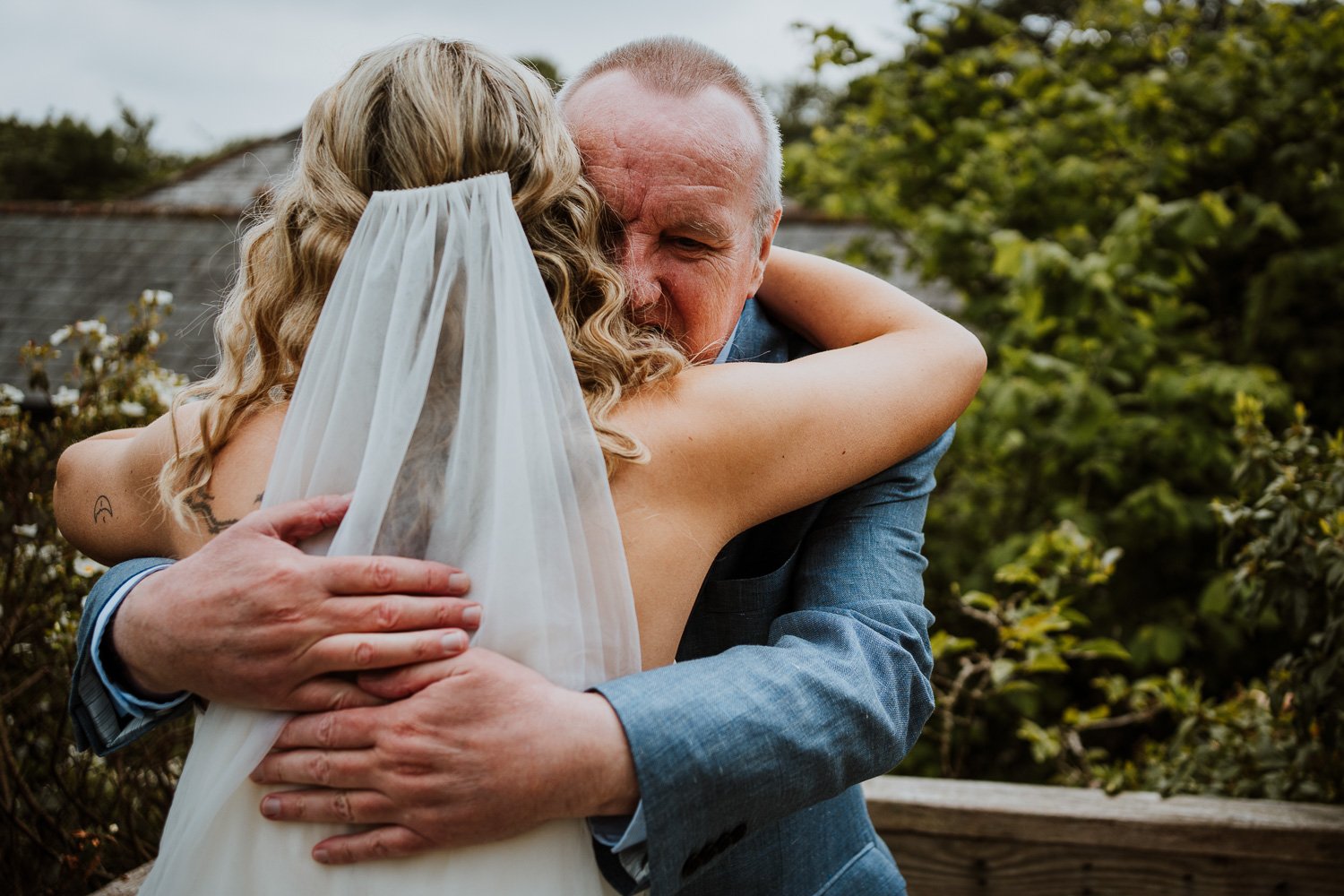 A bride with long curly blonde hair wearing a veil hugs an older man dressed in a blue suit, embracing each other tightly outdoors amidst greenery and a cloudy sky.