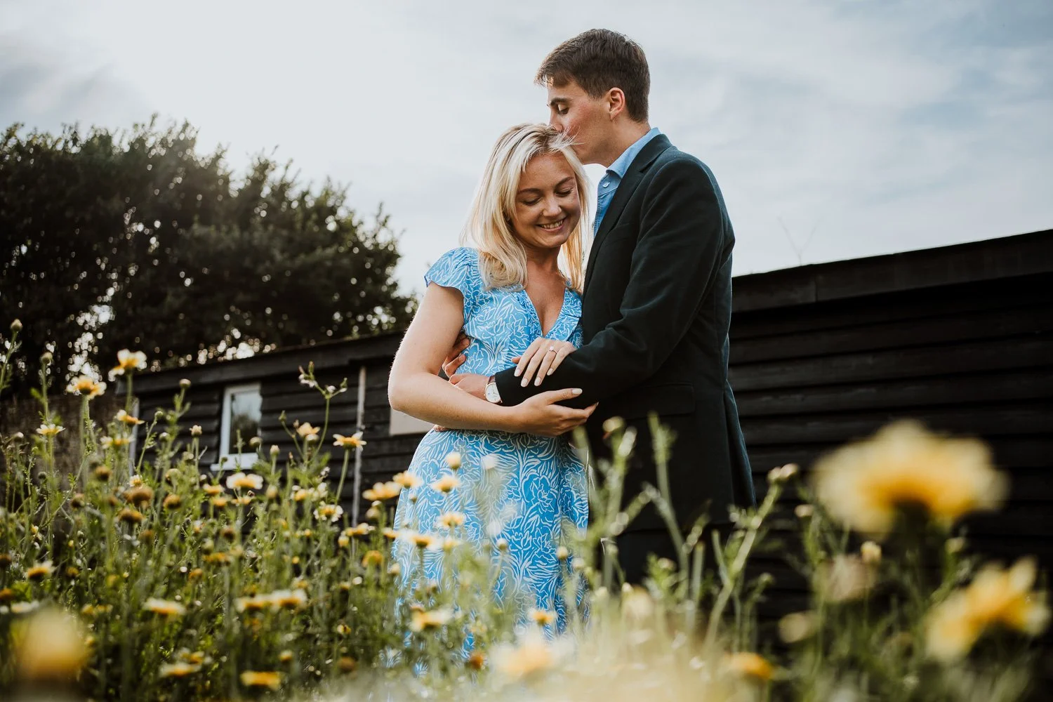 A young couple standing in a flower field, with the man kissing the woman's forehead and both smiling, during daytime. Couple Portraits taken during a Proposal Photography Session