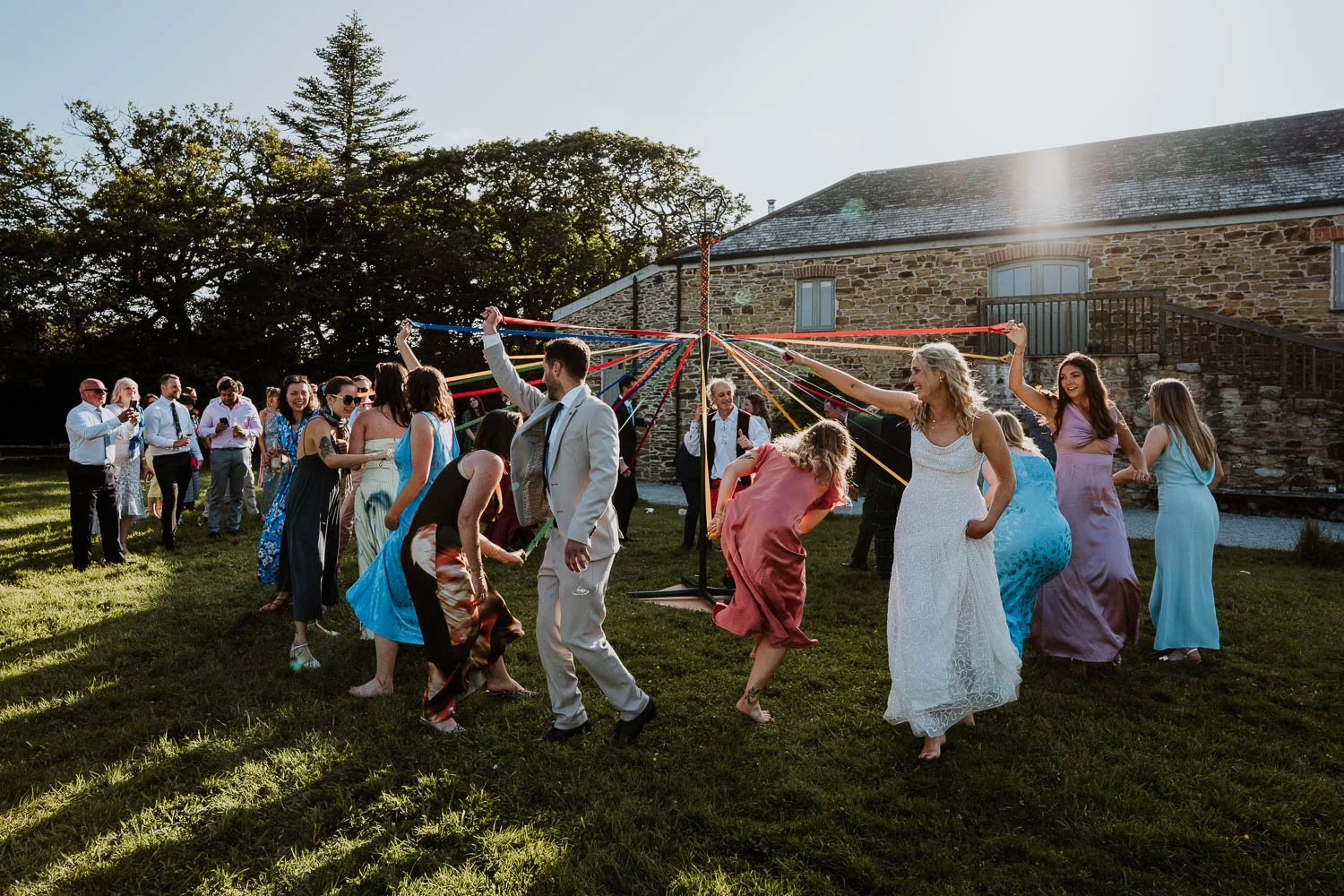 People dancing in a circle at an outdoor wedding reception, with colorful ribbons attached to a central pole and a stone building in the background