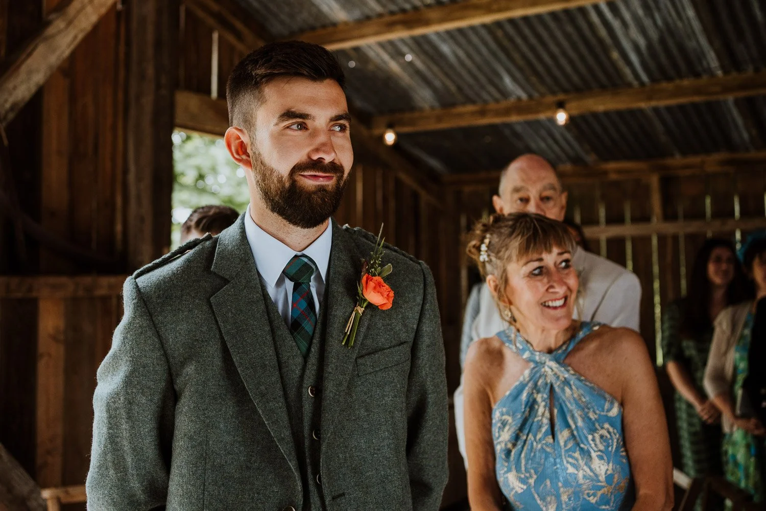 A bearded man in a gray suit and tartan tie stands at a wedding ceremony, smiling. Behind him are an older woman in a blue dress with a floral pattern, and a man in a light-colored shirt. The setting is a rustic wooden barn, with sunlight filtering t