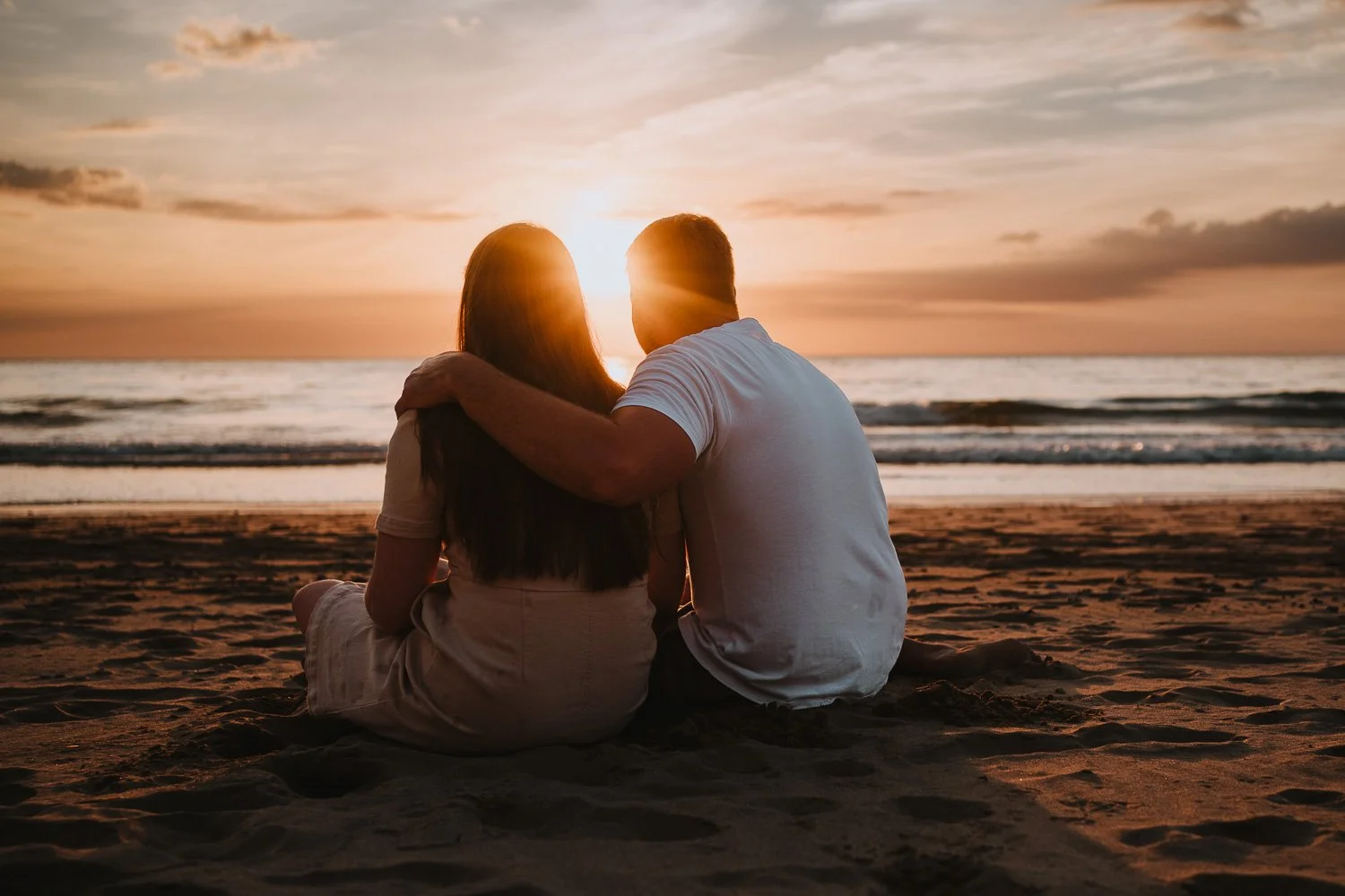 Couple sitting on the beach at sunset, embracing each other and watching the ocean. Couple portrait taken during an engagement / pre wedding photo session