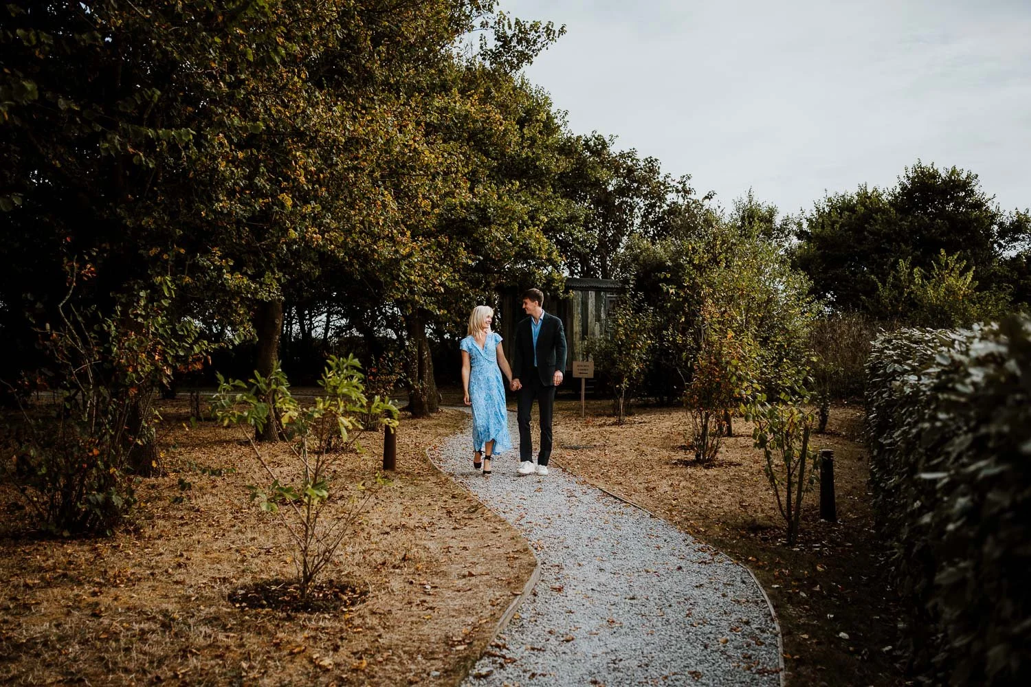 A couple walking hand in hand on a winding gravel path through a garden with trees and bushes, during daytime. Natural Couple Portraits taken during a Proposal Photography Session
