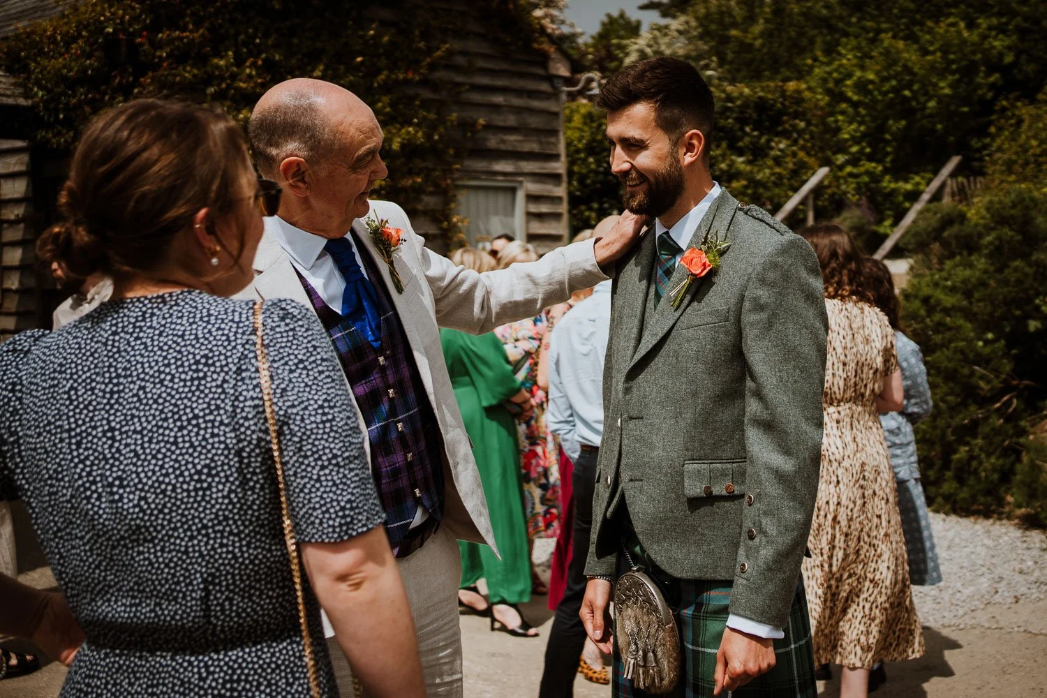Group of people at an outdoor wedding celebration, including a man in a gray kilt and a woman in a patterned dress, with others in colorful attire in the background.