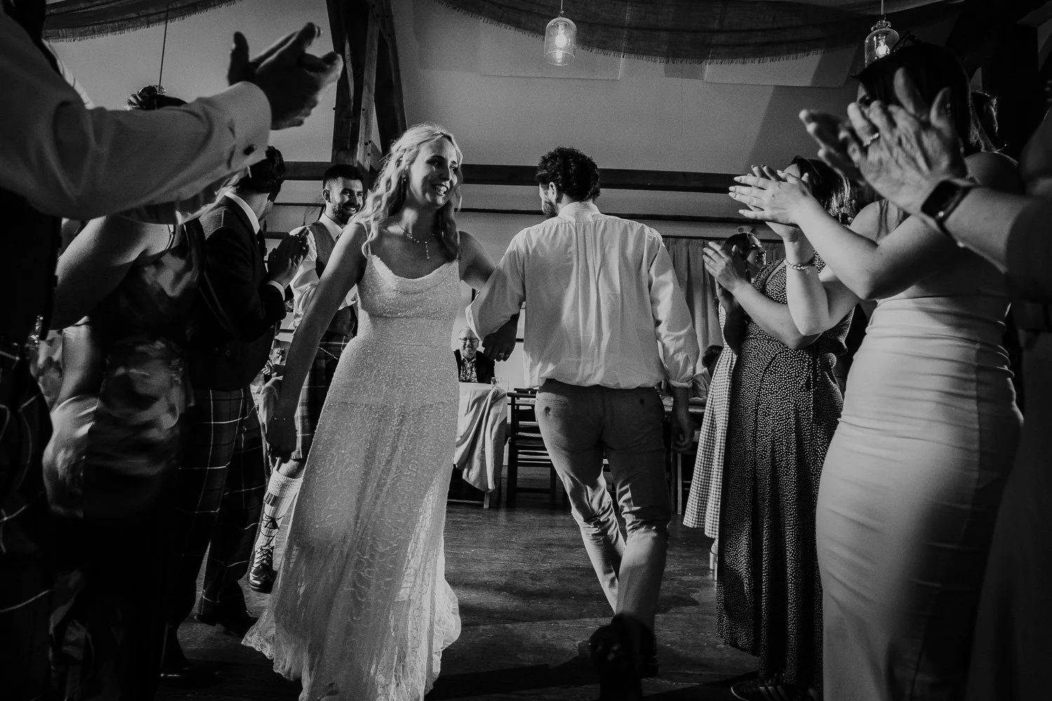 A black-and-white photo of a bride and groom dancing amidst guests at a wedding reception, with guests clapping and celebrating around them. Ceilidh dance fun captured during wedding reception. Black and White Photo