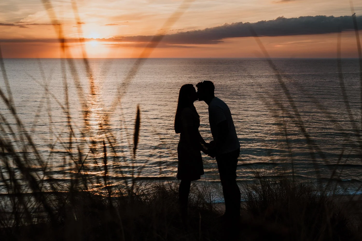 A silhouette of a couple holding hands and about to kiss on a beach at sunset, with the ocean and sky in the background, and wind-blown grass in the foreground. Playful, natural couple portraits taken during pre wedding photo / engagement photo sessi