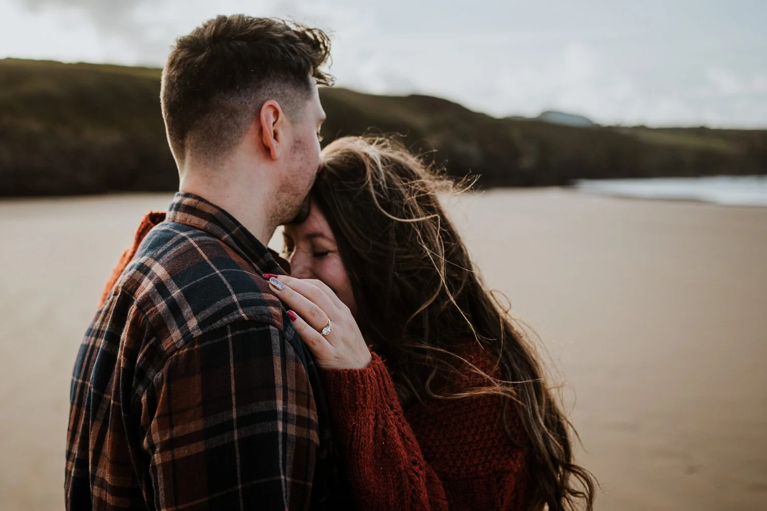 A couple embracing on a beach, with the woman holding the man's shoulder and smiling with her eyes closed, and the man gently resting his forehead on her head, against a backdrop of sand and rocky cliffs. Engagement Photo 