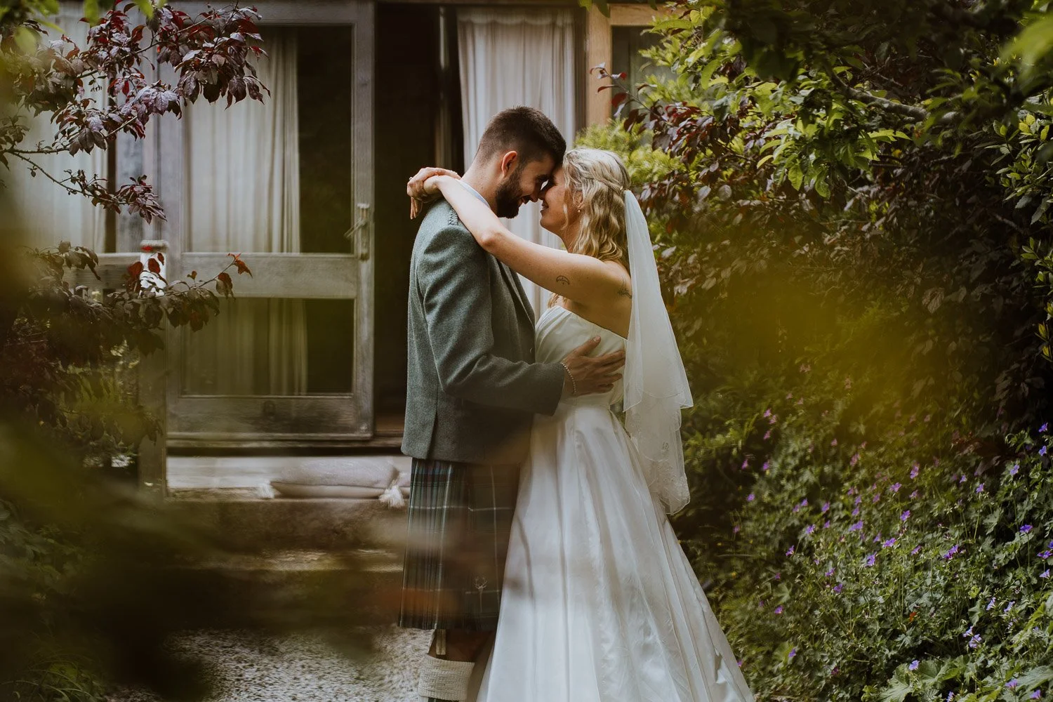 A couple on their wedding day sharing an intimate moment outdoors, surrounded by greenery and flowers, with the bride in a white gown and veil and the groom in a grey suit and kilt. Natural Wedding Couple Portrait