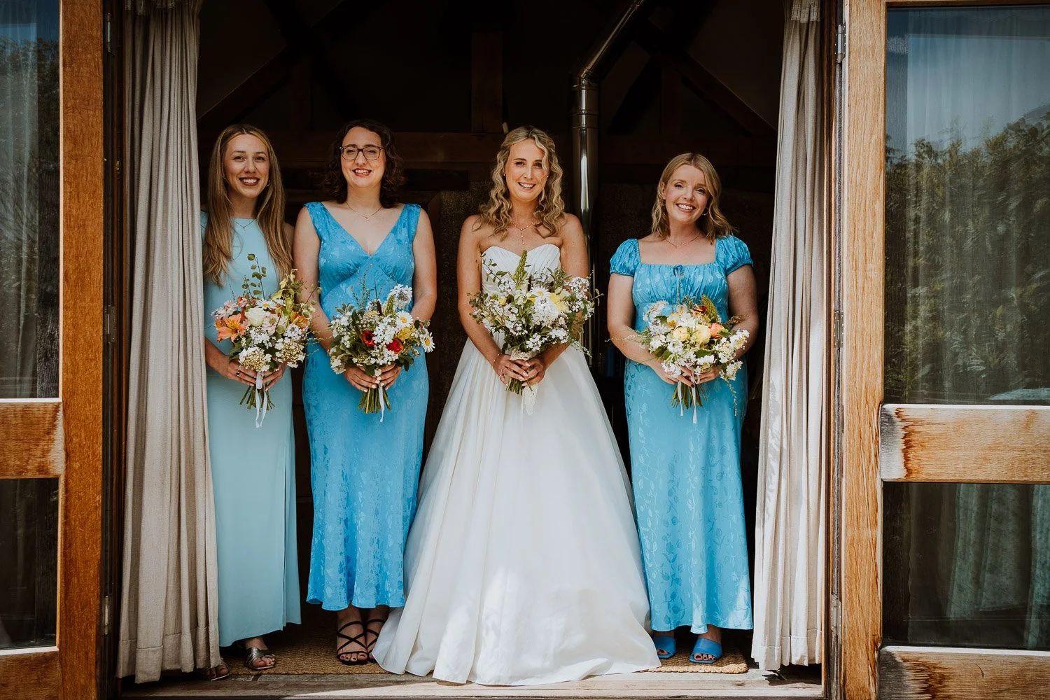 A bride and four bridesmaids standing in a doorway, holding bouquets, dressed in white and blue gowns.
