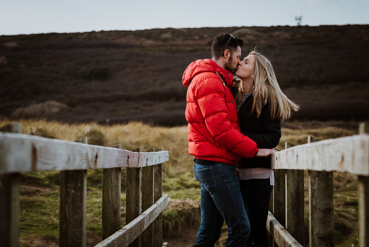 A man and woman sharing a kiss on a wooden bridge in a rural outdoor setting, with hills in the background during late afternoon or early evening. Couple Portrait taken during Engagement Photo Session