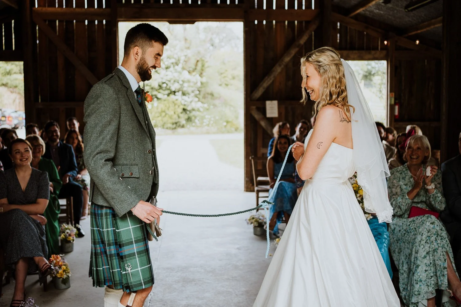 A bride and groom during their wedding ceremony in a rustic wedding barn. A knot-tying wedding ceremony, or "handfasting" ceremony where bride and groom tying a knot in a rope as a symbol for their love