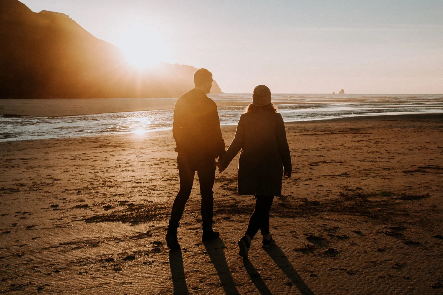 A couple holding hands walks along the beach at sunset, with the sun low on the horizon and casting long shadows. Couple portrait taken during an engagement photo session
