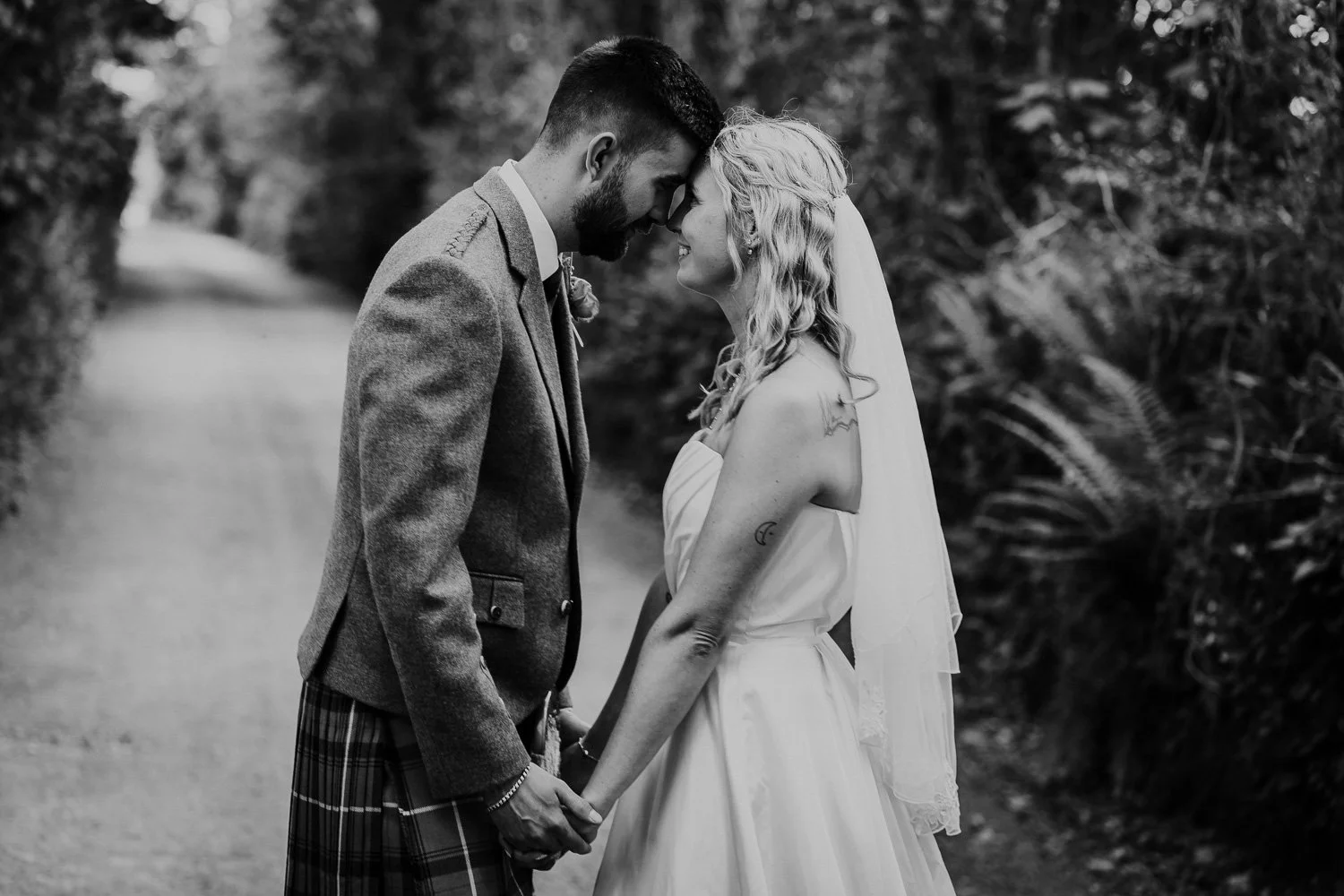 A black and white photograph of a bride and groom standing close and touching foreheads in a natural outdoor setting, holding hands, with trees in the background. Natural Wedding Couple Portrait