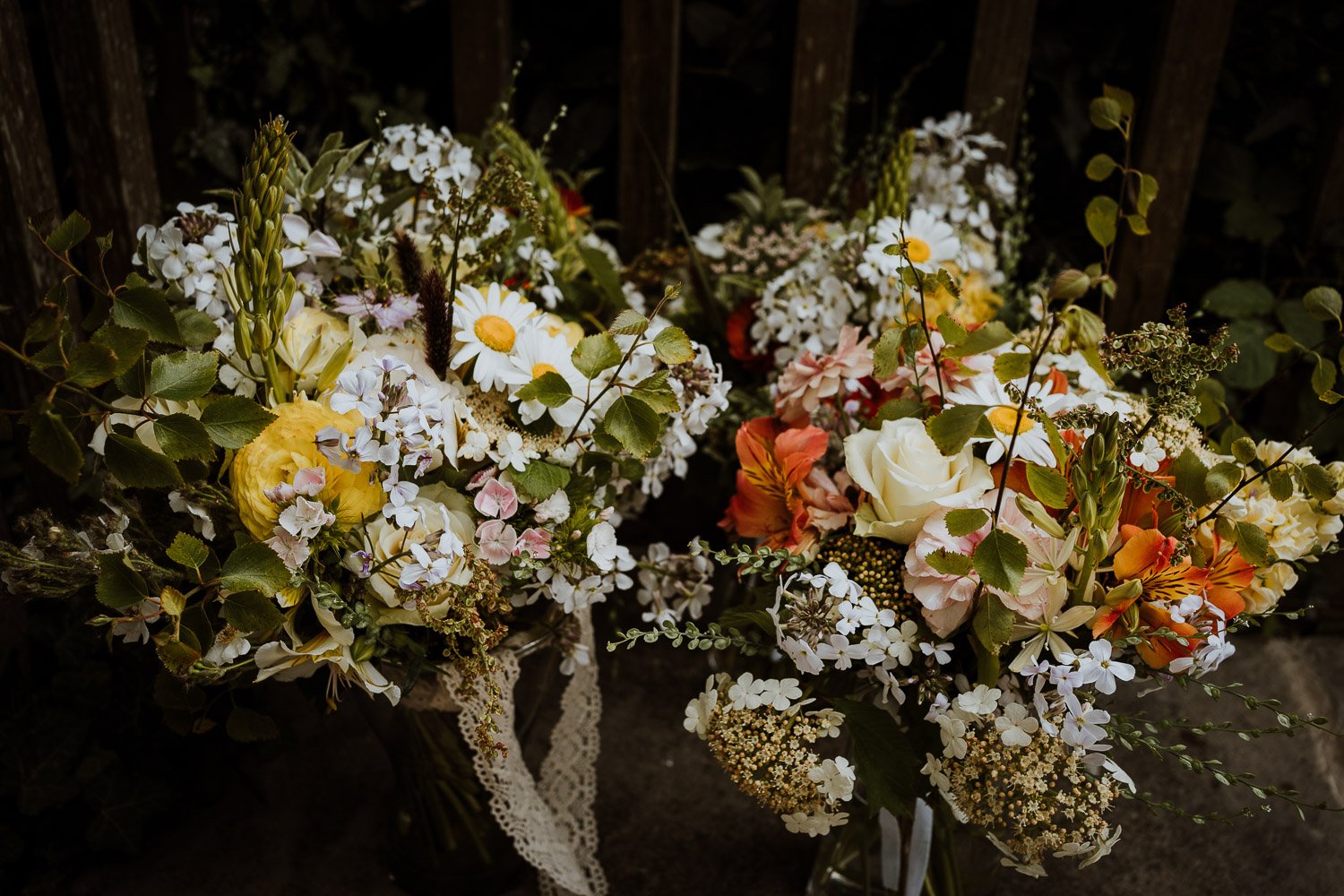 Two bridal bouquets of mixed seasonal flowers including daisies, roses, hydrangeas, and other small flowers, arranged with greenery, placed on a dark surface near a wooden fence.