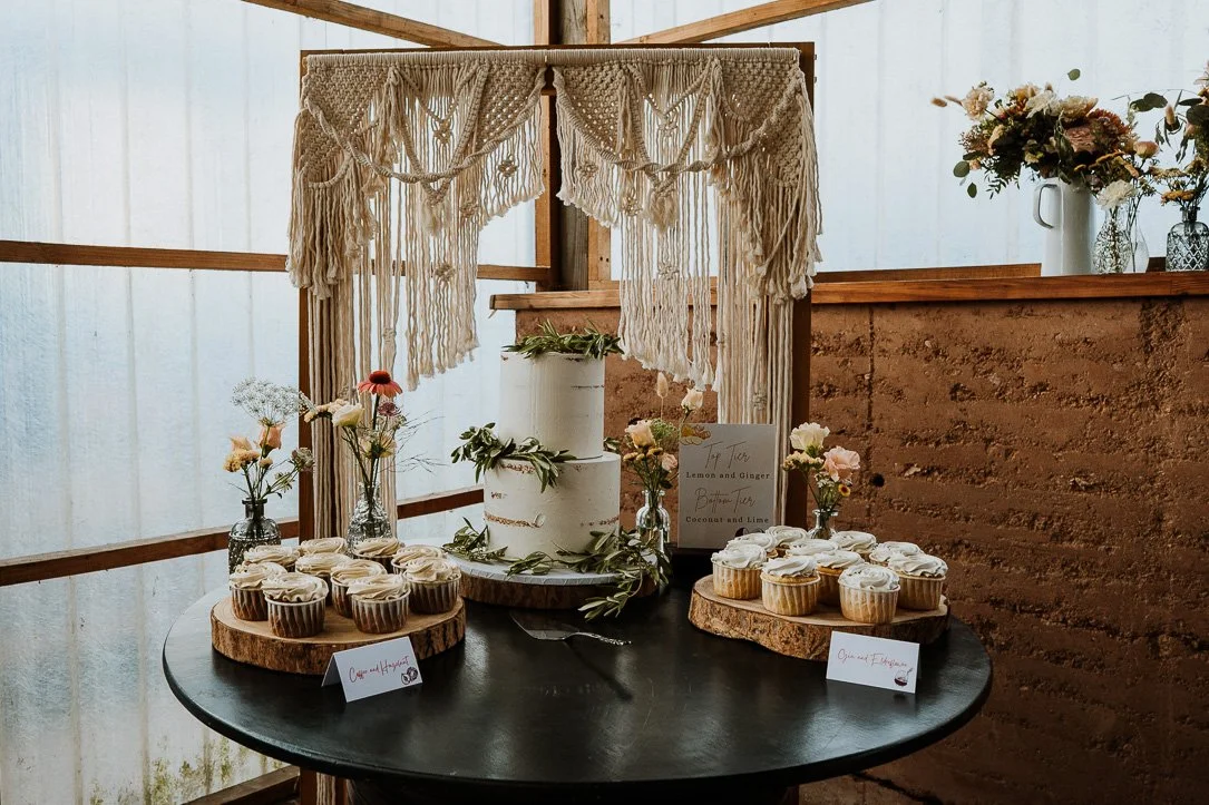 A dessert table with cupcakes, a wedding cake, flowers, and a sign in a rustic setting 