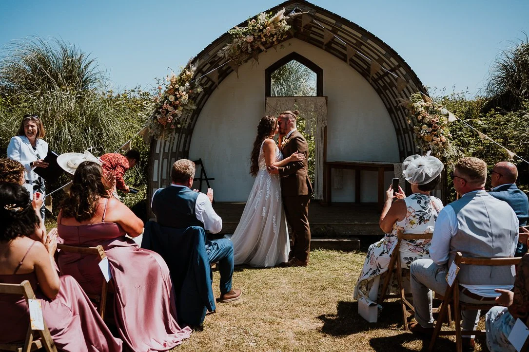 A bride and groom share a kiss during their outdoor wedding ceremony on a sunny day, surrounded by guests seated on wooden chairs, with a decorative floral arch behind them.