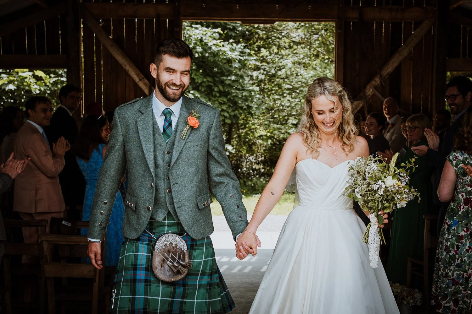 A newlywed couple holding hands and smiling during their exit from the wedding ceremony inside a rustic wooden barn with guests applauding in the background.