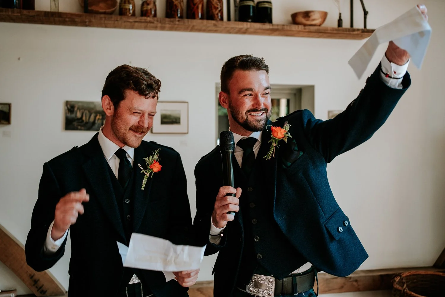Two men dressed in dark suits with orange boutonnieres, one holding a microphone and the other holding a paper, smiling and celebrating indoors.