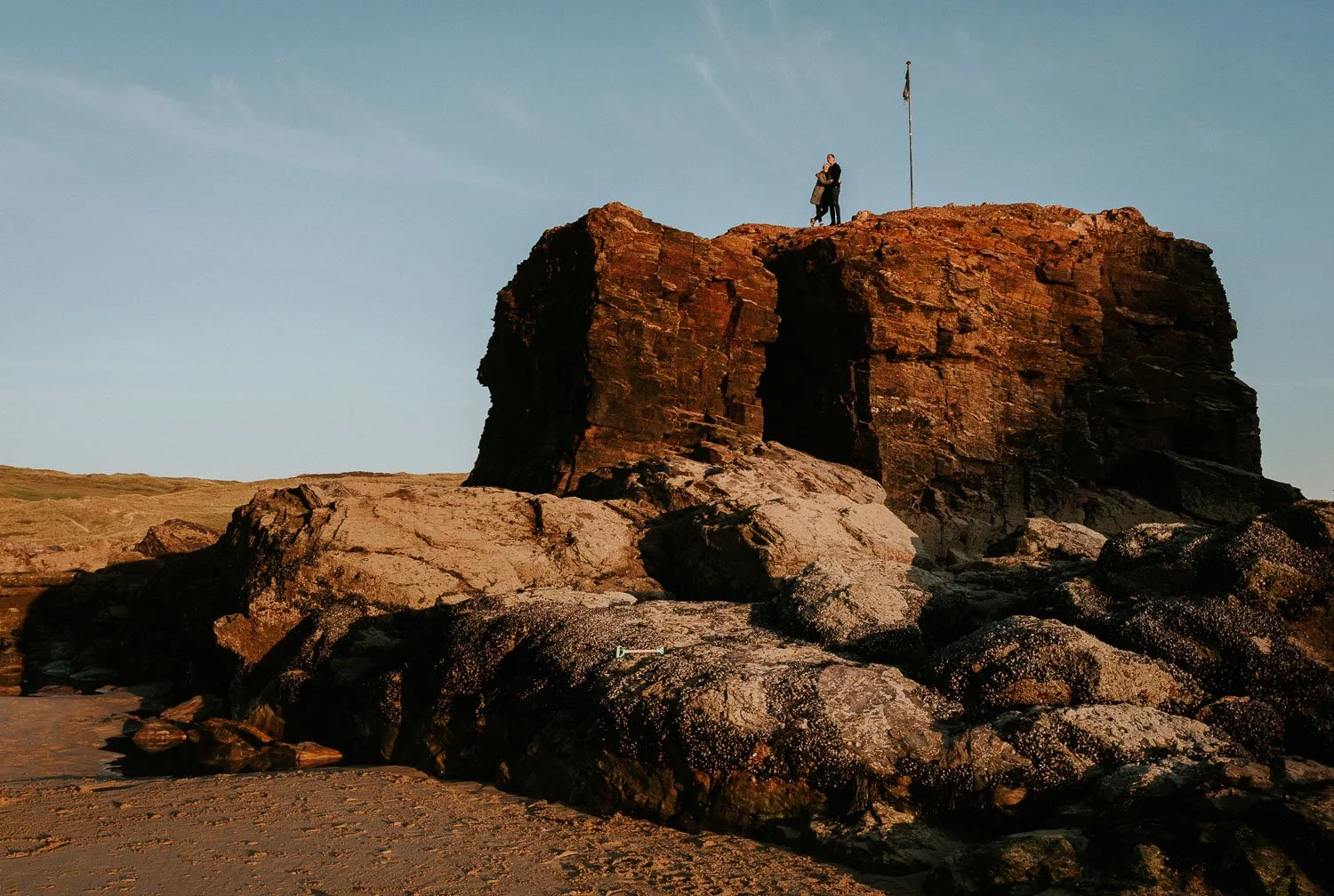Two people standing on top of a large rocky formation on the shoreline under a clear sky at sunset. Couple portrait taken during an engagement photo session