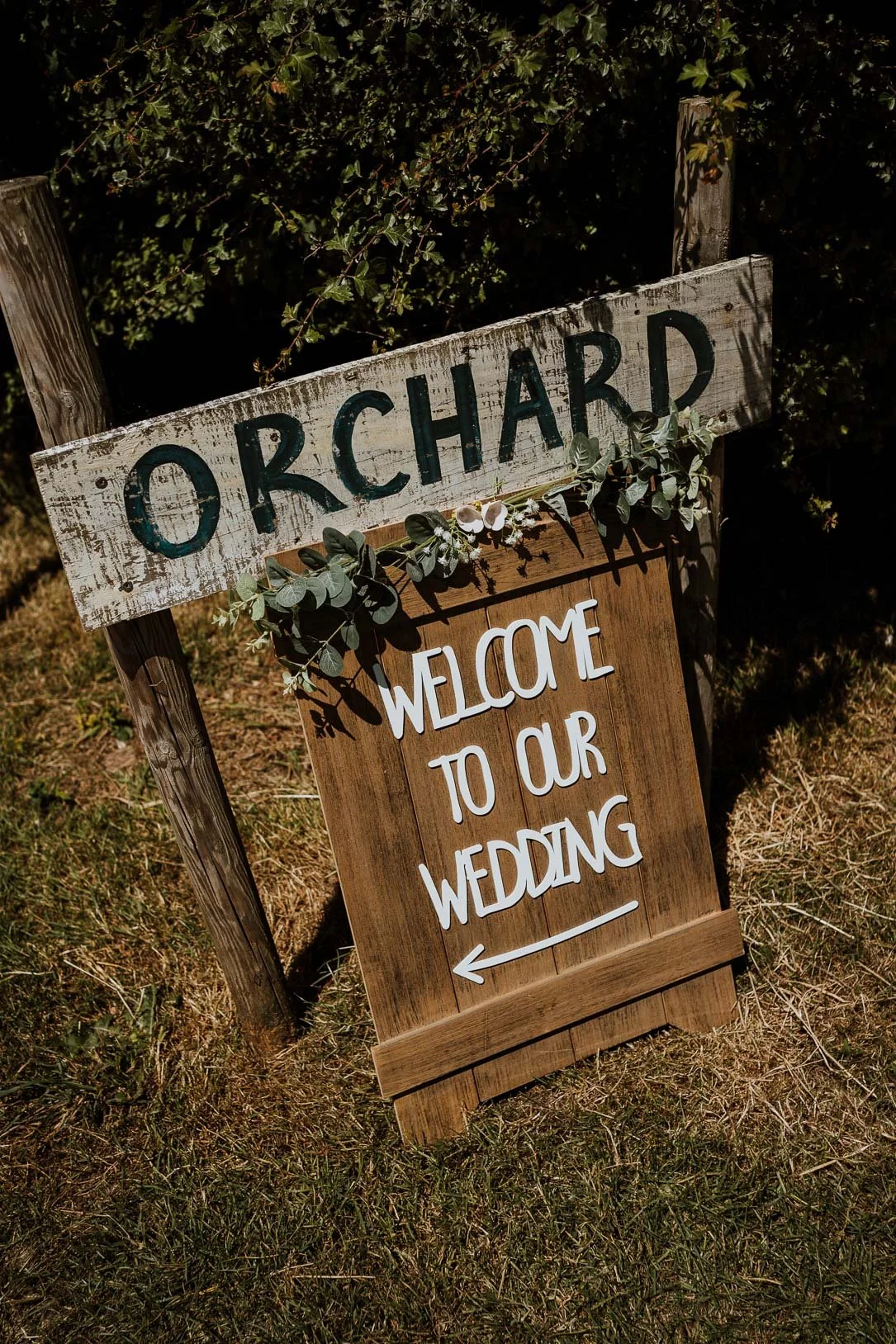 Wooden wedding sign reading 'Welcome to our wedding' with an arrow pointing left, decorated with eucalyptus leaves and flowers, standing outdoors on grass.