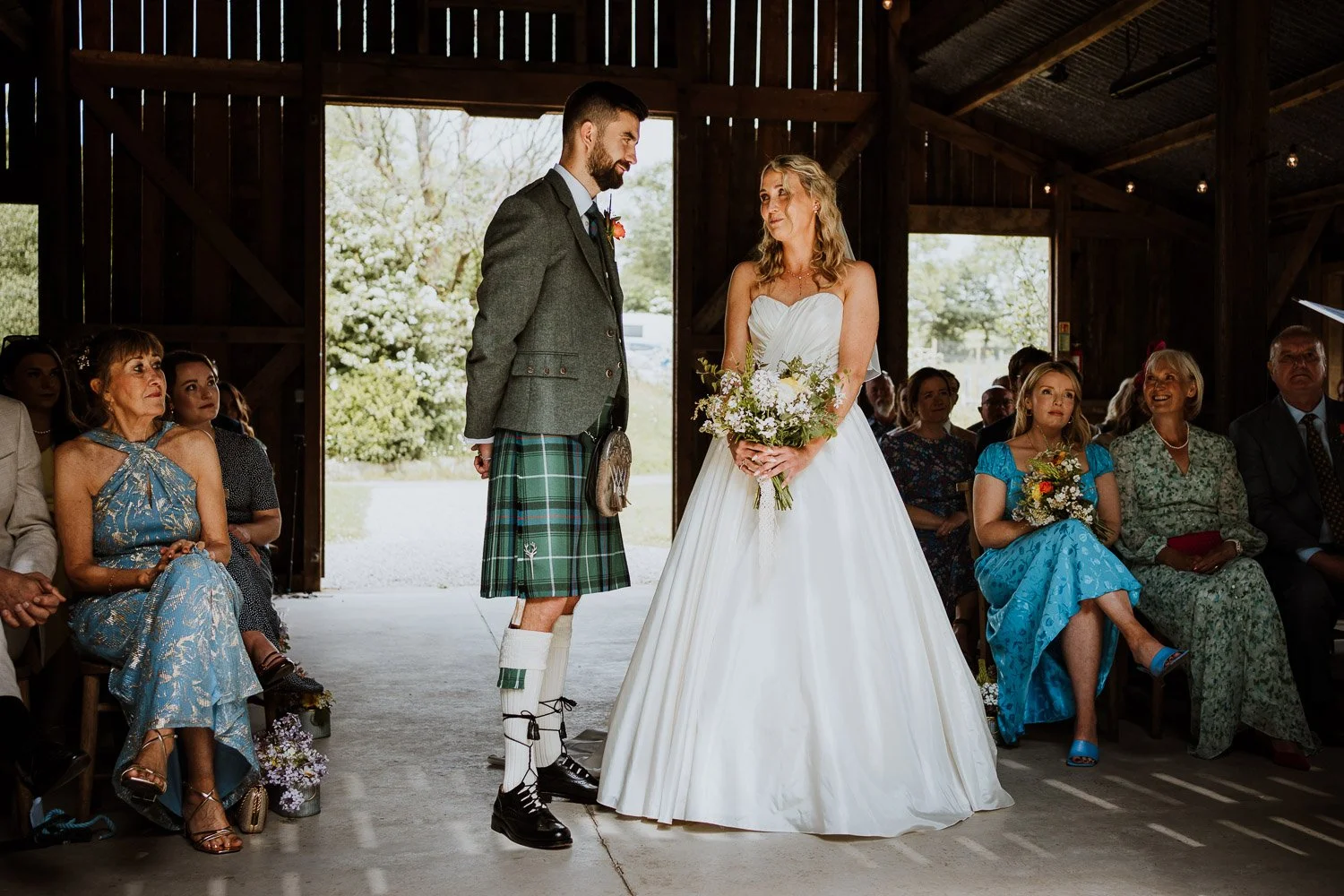 A wedding ceremony inside a rustic barn, with a bride in a white gown holding a bouquet and a groom wearing traditional Scottish attire, including a kilt and sporran, standing face to face. Guests are seated on either side, watching the ceremony.