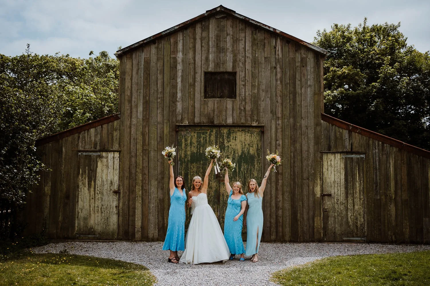 Bridesmaids in blue bridesmaid dresses and bride in front of a rustic wooden barn, holding bouquets and raising their arms joyfully. Group Photos