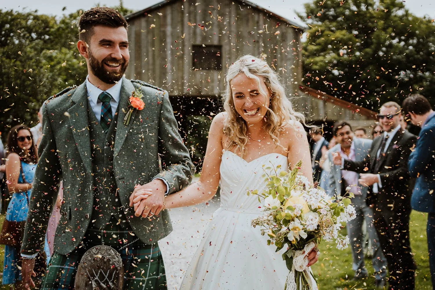 A newlywed couple, a man in a kilt and a woman in a white wedding dress, walking hand in hand outdoors as confetti falls around them, surrounded by wedding guests, with a rustic barn in the background. Nancarrow Farm Wedding