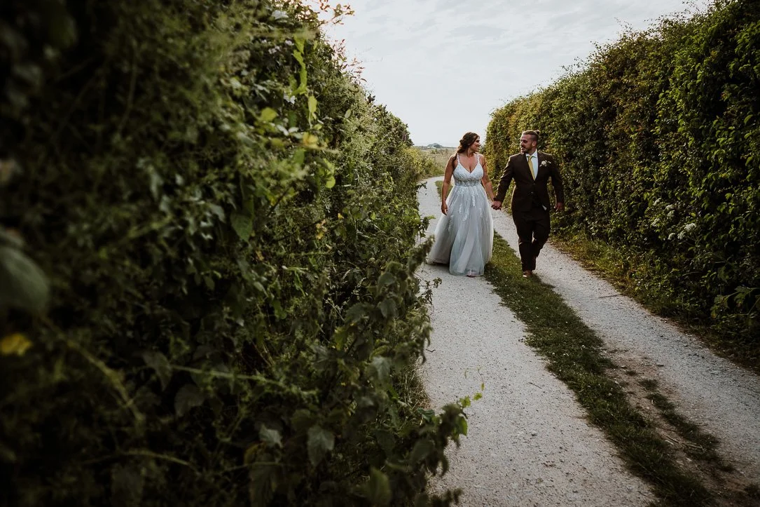 A bride and groom walking hand in hand down a dirt path surrounded by green hedges, dressed in wedding attire. Natural Wedding Couple Portrait Photo