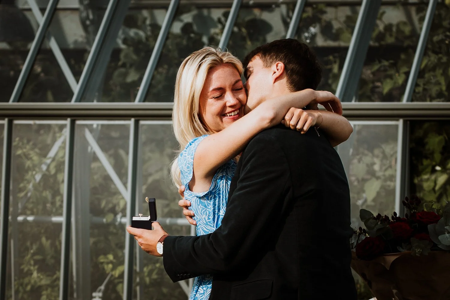 A woman with blonde hair in a blue dress is hugging a man in a black suit, who is holding an open ring box. They are smiling and appear very happy, possibly during a proposal or celebration. Natural Proposal Photography