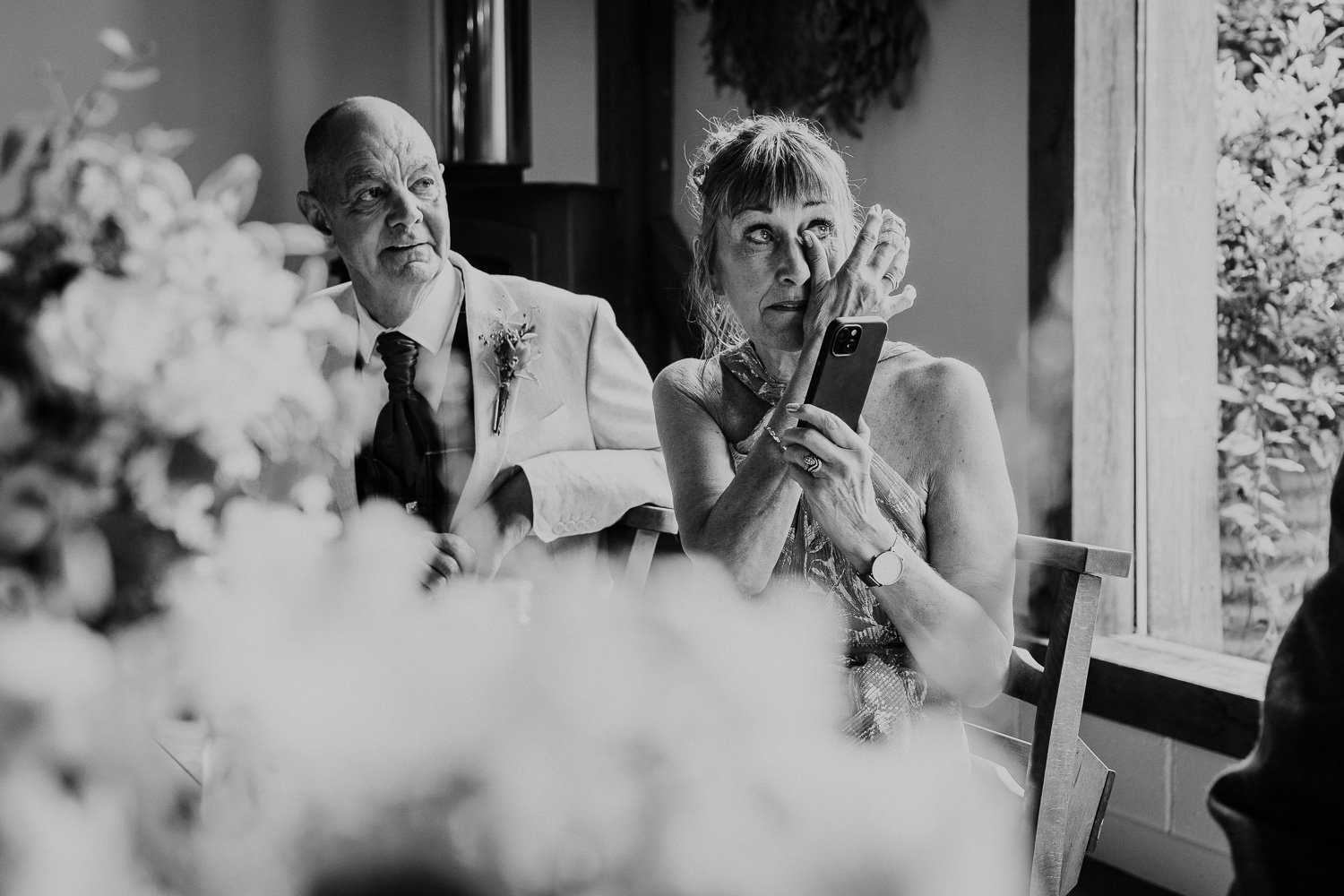 A black-and-white photo of a woman wiping tears from her eyes while holding a smartphone, seated next to an older man wearing a suit and tie at a social gathering or event.