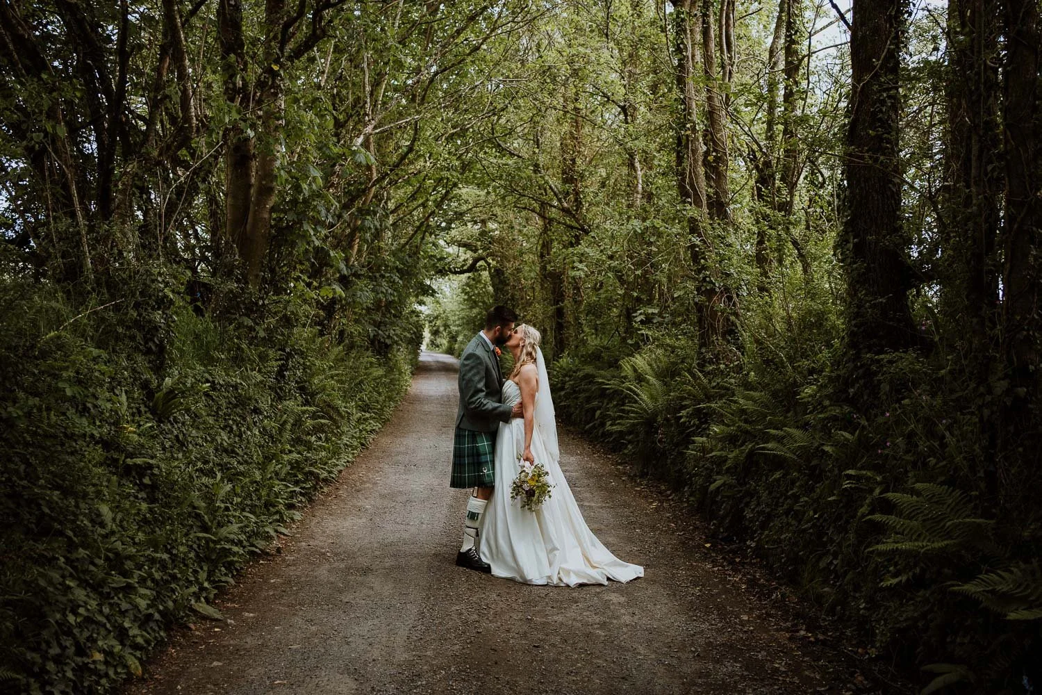 A bride and groom sharing a kiss on a forested dirt path, surrounded by lush green trees and ferns. Woodland Couple Portrait