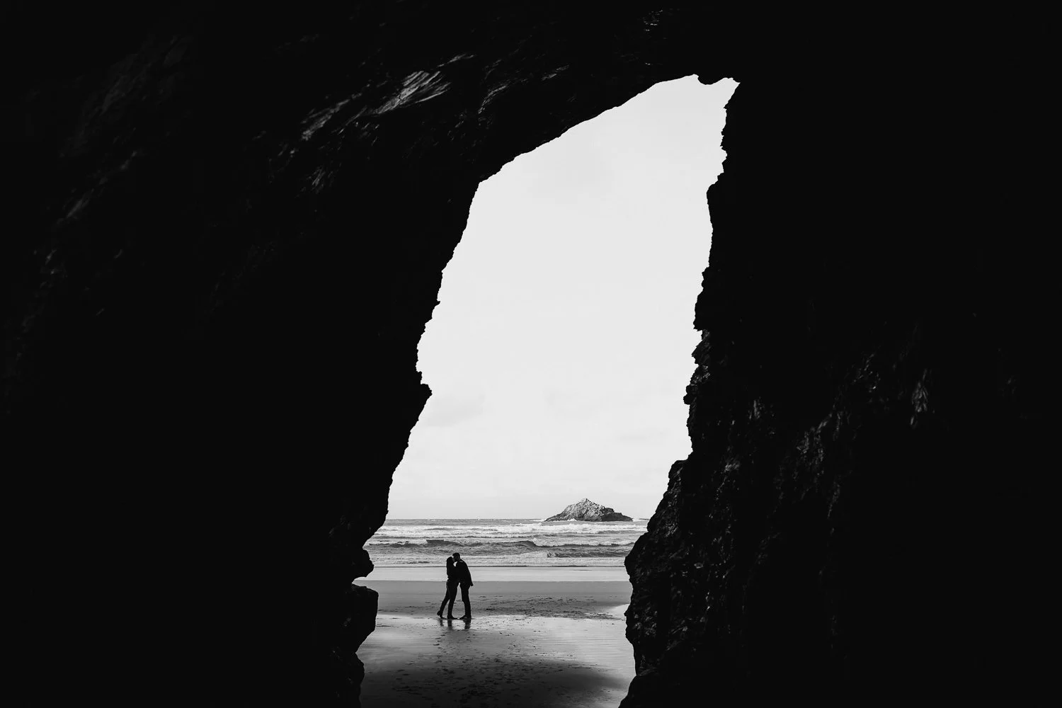Couple kissing on beach seen through a large cave opening with rocks and ocean in background