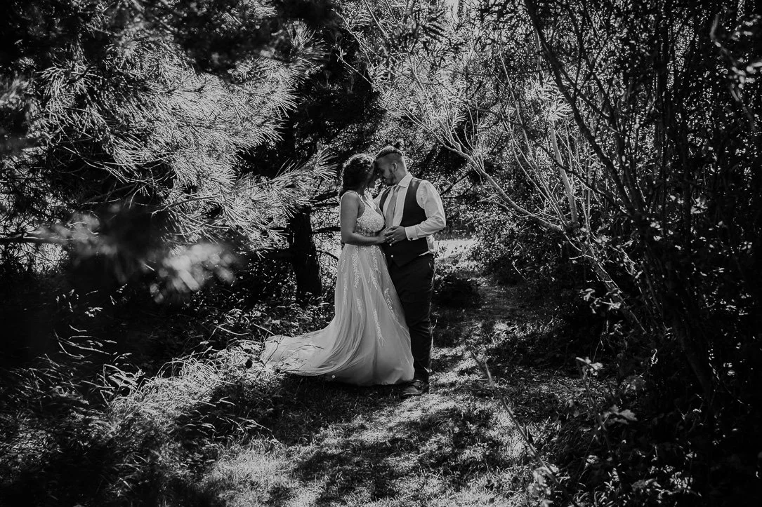 Black and white photo of a couple in wedding attire standing close together in a wooded outdoor setting, holding hands and touching foreheads under trees. Natural Couple Portrait