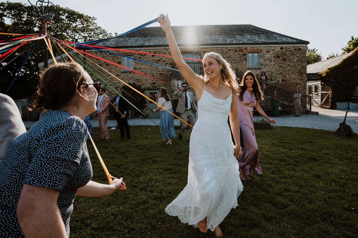 A bride in a white wedding dress smiling and dancing during a celebration outside a stone building, surrounded by guests in colorful dresses and suits, with festive ribbons attached to a central pole.
