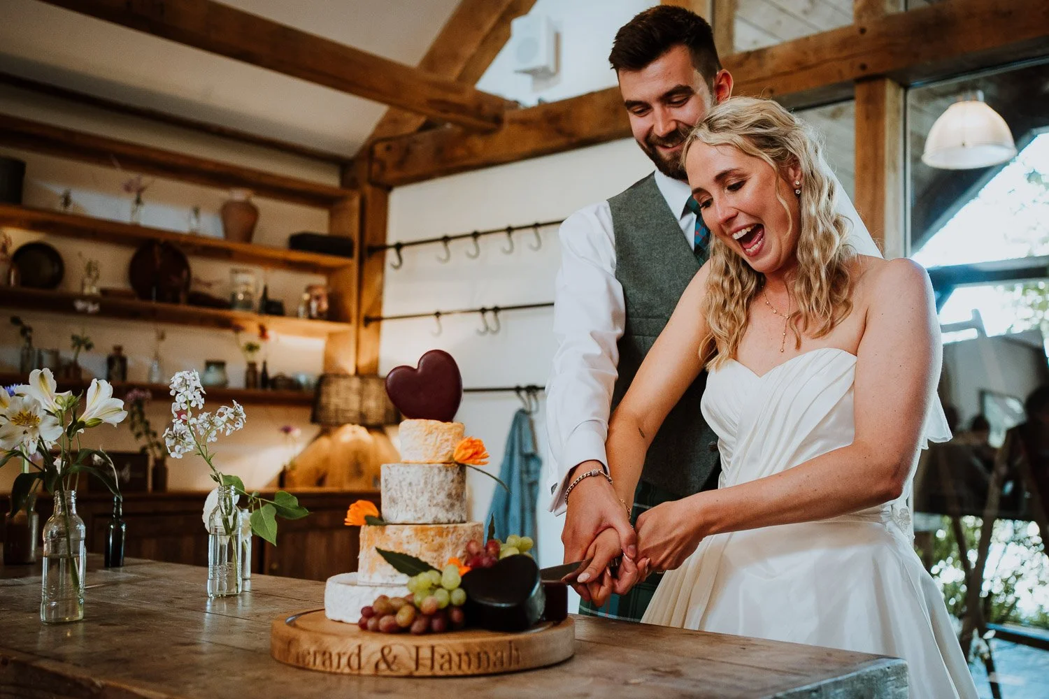 A newlywed couple happily cuts their wedding cake together at a rustic indoor venue with wooden decor and shelves in the background.