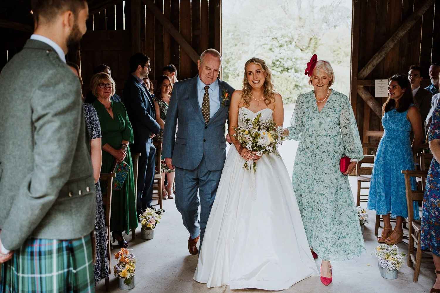 A bride in a white wedding dress walking down the aisle accompanied by her parents in a rustic wooden barn.