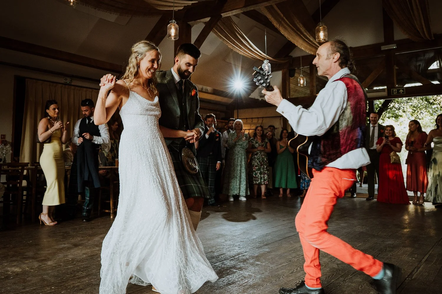 Wedding reception with bride and groom dancing, surrounded by guests, a musician playing guitar, and decorated with warm lighting.