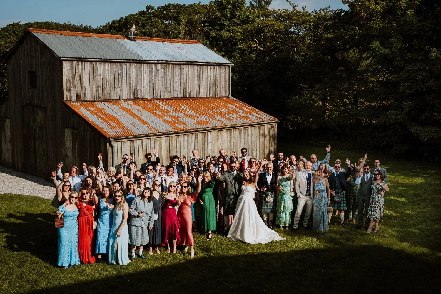 Large group of wedding guests, including bride and groom, posing outdoors in front of a rustic wooden barn, with many smiling and raising their hands.