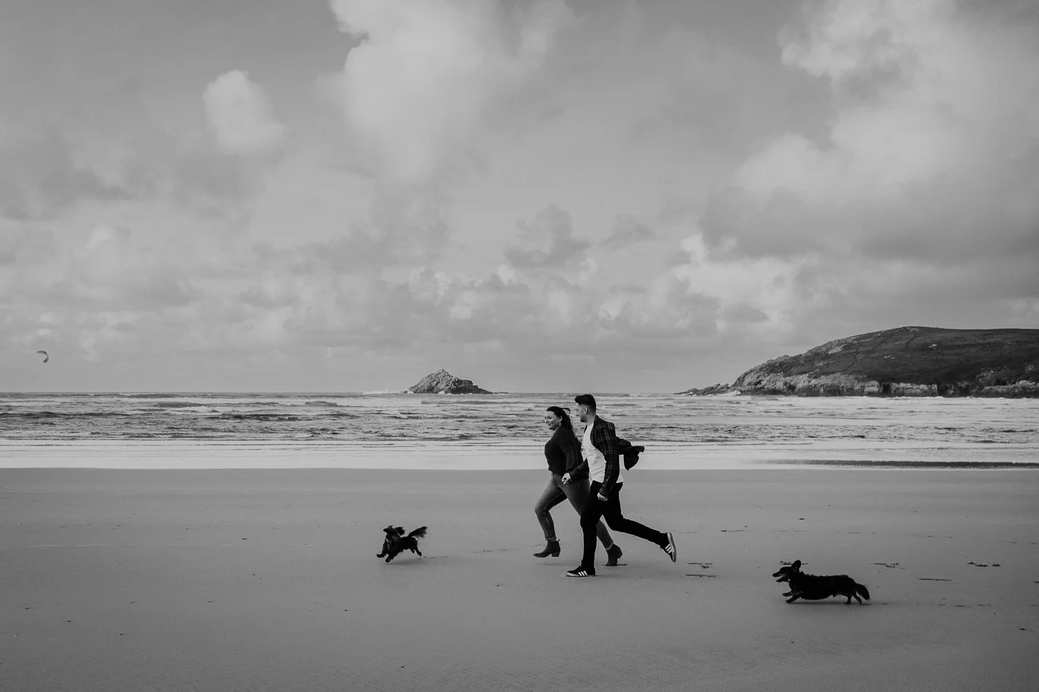 A black and white photo of a couple jogging on the beach with two dogs; the ocean, cloudy sky, and distant hills are in the background. Engagement Photo of couple and their dogs on the beach in Cornwall. Black and White Photo
